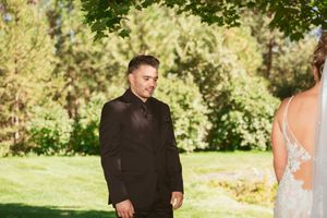 A groom in a black suit stands outdoors on a sunny day with trees and greenery in the background.