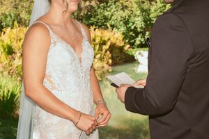 In outdoor wedding ceremony, two formal figures exchange rings during their vows beneath green trees and natural lighting.