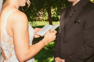 A person in a lace wedding dress reads vows from a phone during an outdoor ceremony while facing someone in a black suit.