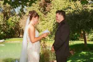 Two newlyweds share a tender moment while exchanging vows in a sun-dappled garden setting with lush greenery.