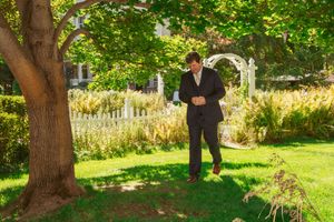 Someone in a dark suit stands beneath a shady tree near a white garden arch on a manicured lawn.