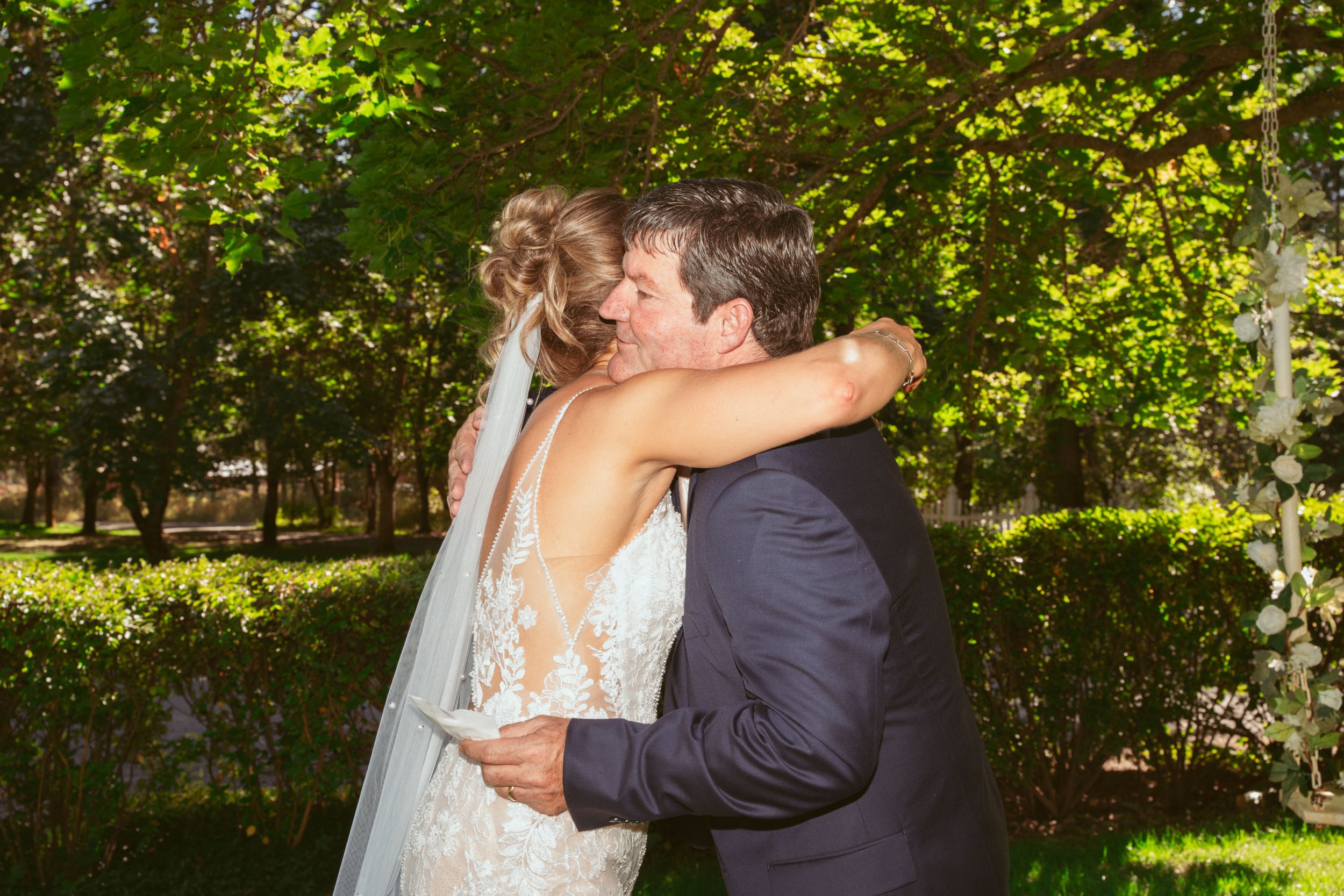 Bride and groom embrace while dancing outdoors beneath green trees at their wedding celebration.