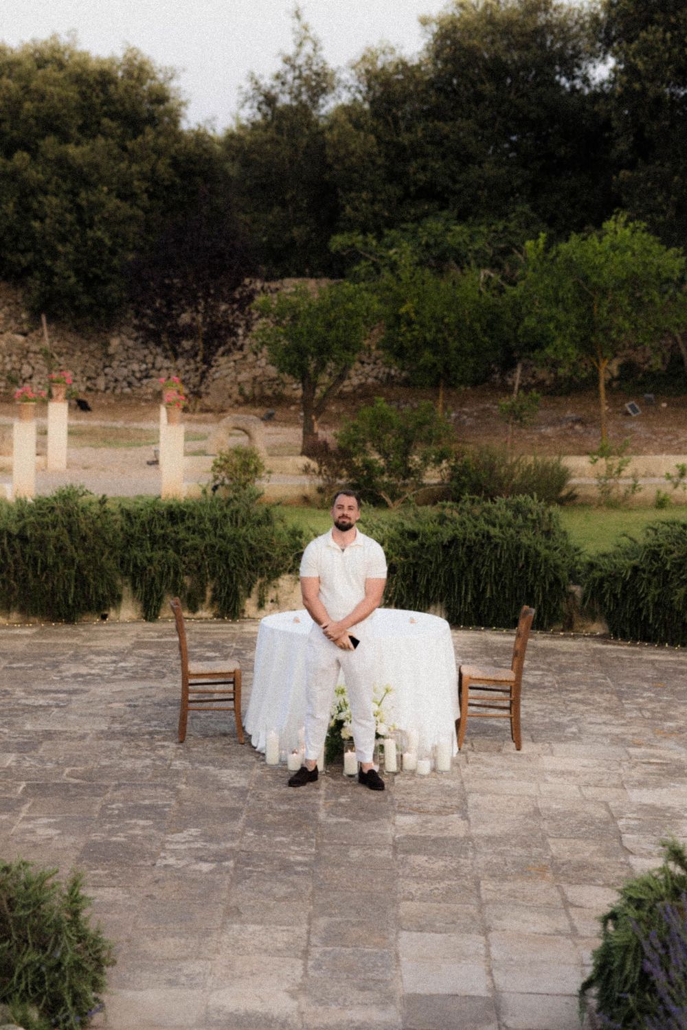 An elegant outdoor table setting with white linens and chairs on a stone patio surrounded by lush greenery and trees.