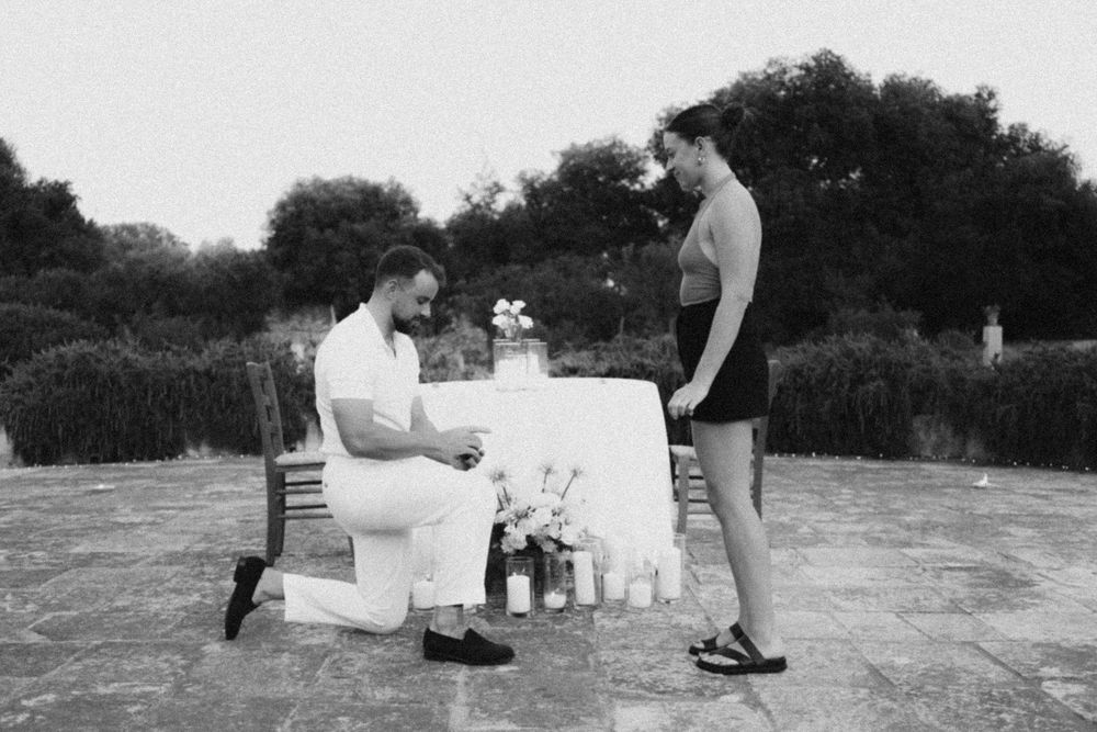 A vintage black and white photo showing two people at a picnic setup outdoors with a blanket and bottles.