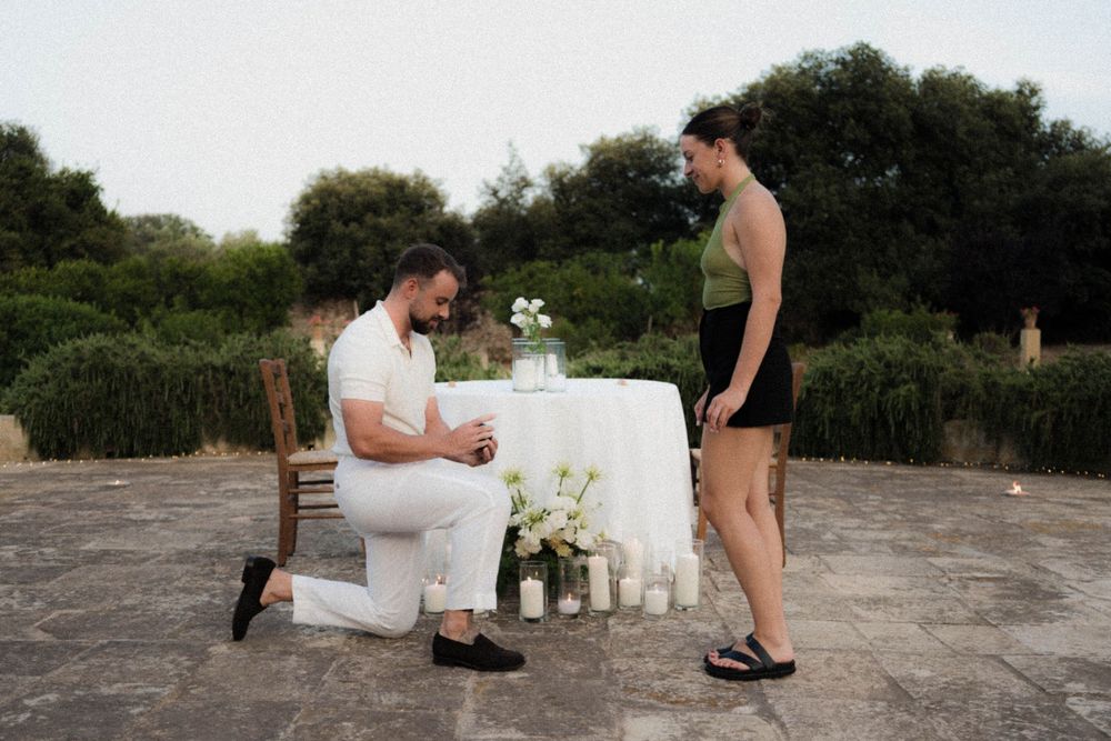 A person proposes on one knee in front of a decorated white table display outdoors on a stone surface.
