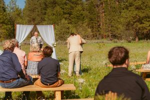 Wedding guests seated on wooden benches in an outdoor meadow with pine forest backdrop.
