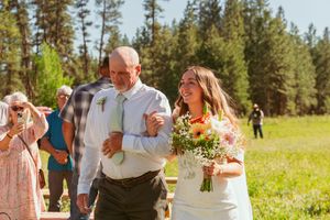 A laughing couple walks arm in arm across a sunlit meadow with pine trees in the background.