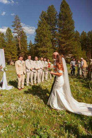 Outdoor wedding ceremony in a mountain meadow with wildflowers and pine trees, featuring wedding party in beige and white.