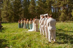Wedding processional line in a sunny mountain field with pine trees in the background wearing beige and white attire.