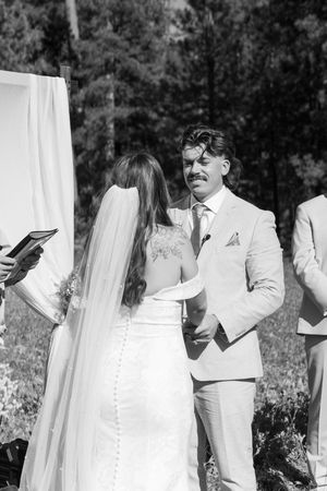 Outdoor wedding ceremony scene with bride in white lace dress and groom in light gray suit exchanging vows.