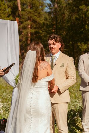 Outdoor wedding ceremony with people in tan and white attire standing beneath sheer white curtains.