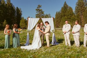 Panoramic view of wedding ceremony in mountain meadow with white draped arch and guests seated on benches.