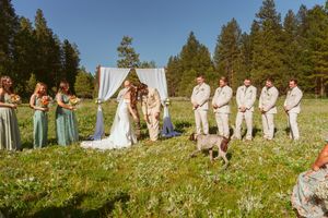 Wedding ceremony group arranged in mountain meadow with white draped arch and evergreen forest backdrop.
