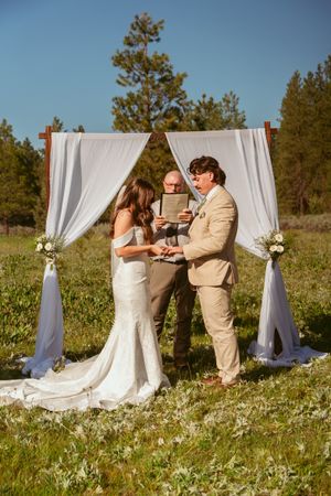 Wedding ceremony outdoors with white fabric arch in meadow of wildflowers and pine trees on sunny day.