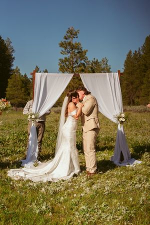 Intimate wedding moment under white fabric arch in sunlit meadow with pine forest backdrop.