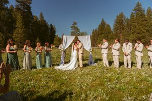 Wedding party lined up in meadow with white arch and evergreen forest in background on sunny afternoon.