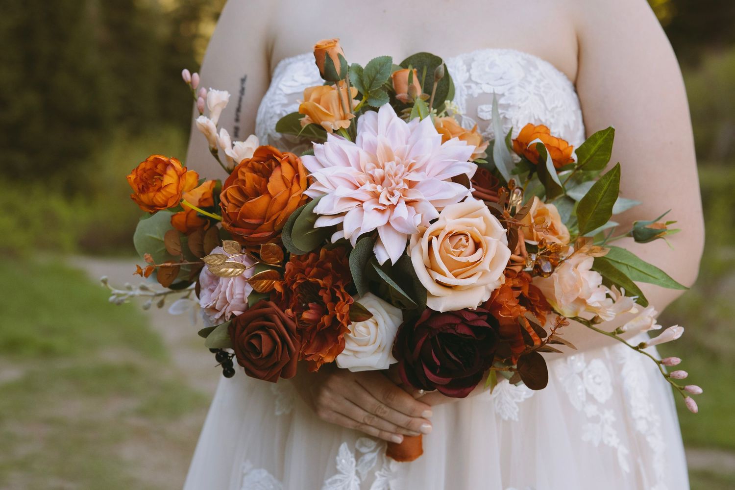 Close-up of autumn-themed bridal bouquet with peach dahlias, orange roses and greenery against a white wedding dress.