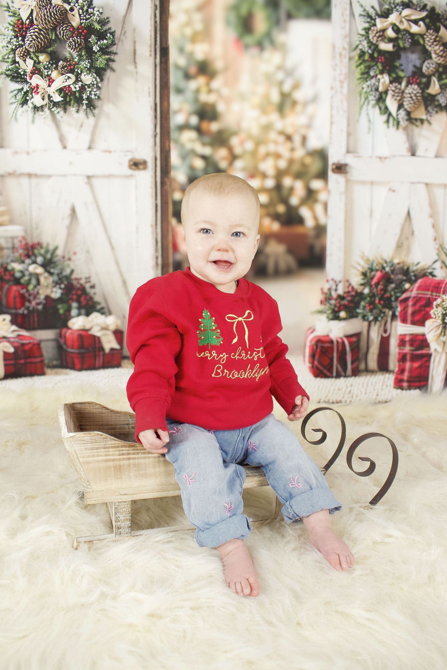 Baby wearing festive red sweater sitting in wooden sleigh with Christmas decorations.