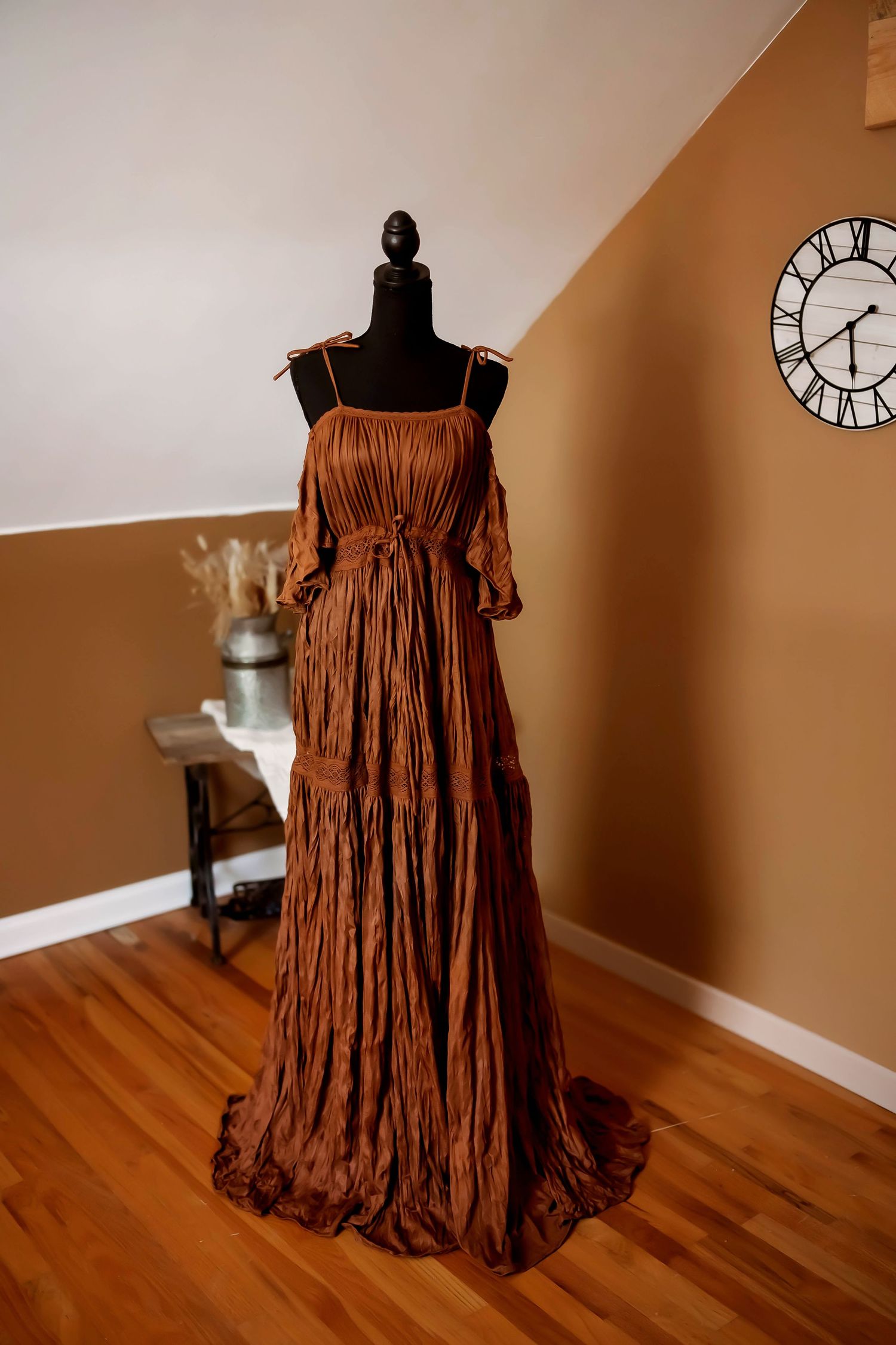 A floral maternity gown with long train displayed on black mannequin in peachy brown tones on wooden floor.