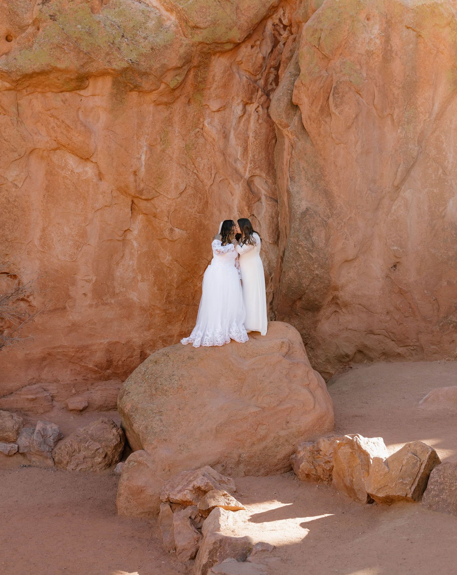 Two people in white wedding dresses stand together on a red rock formation in a desert canyon.