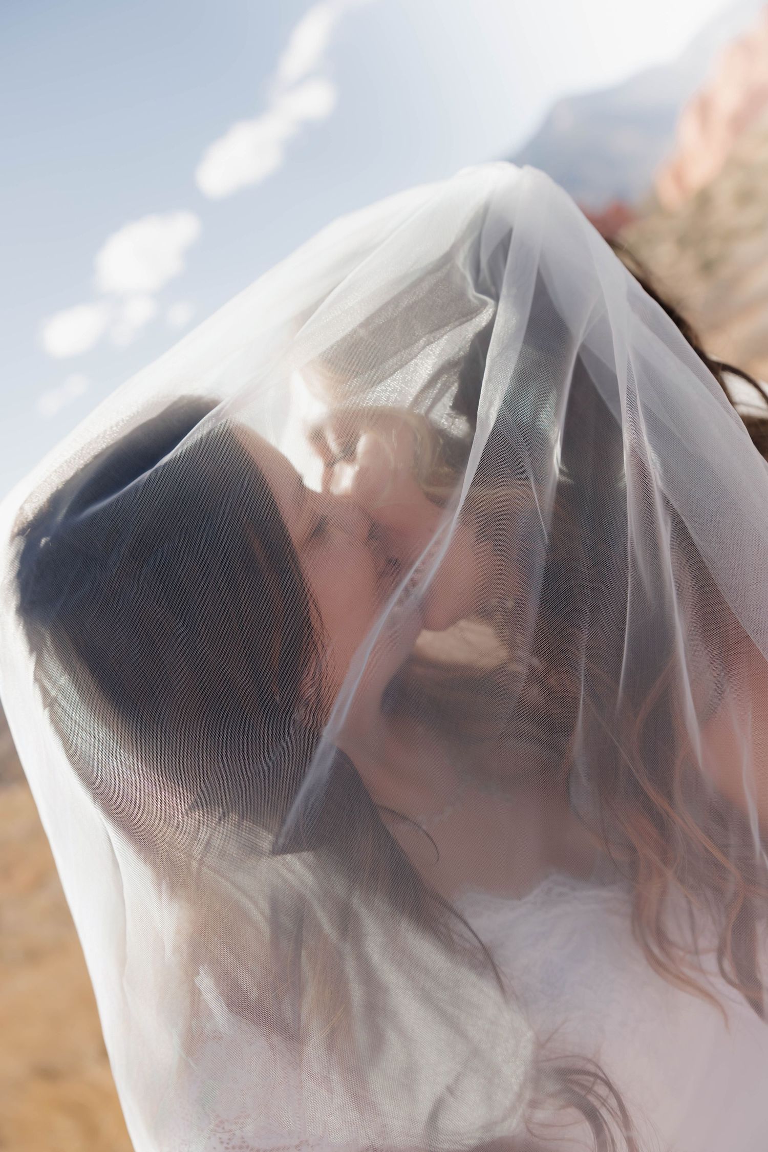 A delicate bridal veil flows in the breeze against a soft blue sky and desert mountain backdrop.