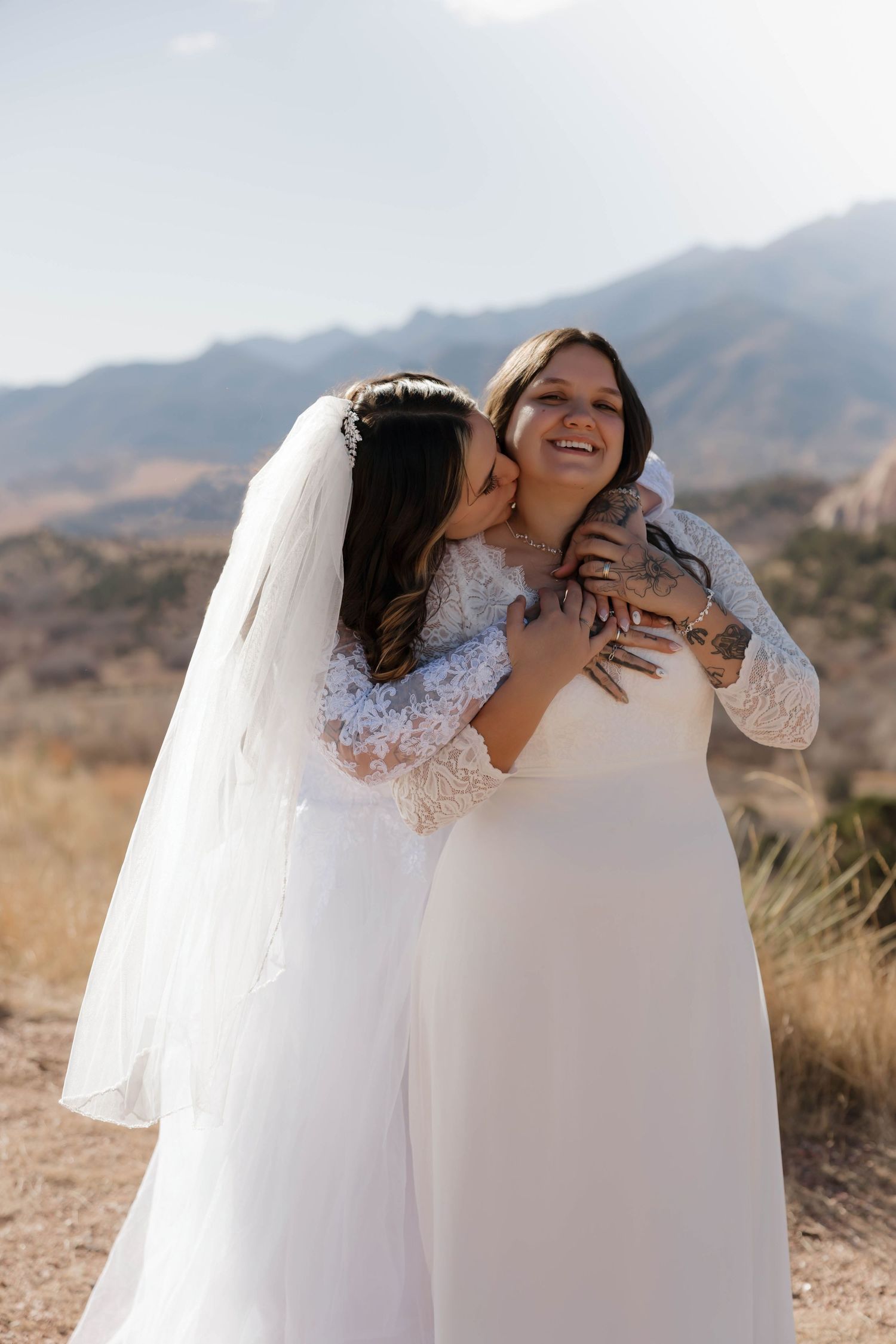 Two people in white dresses embrace joyfully outdoors with mountains and desert landscape in the background.