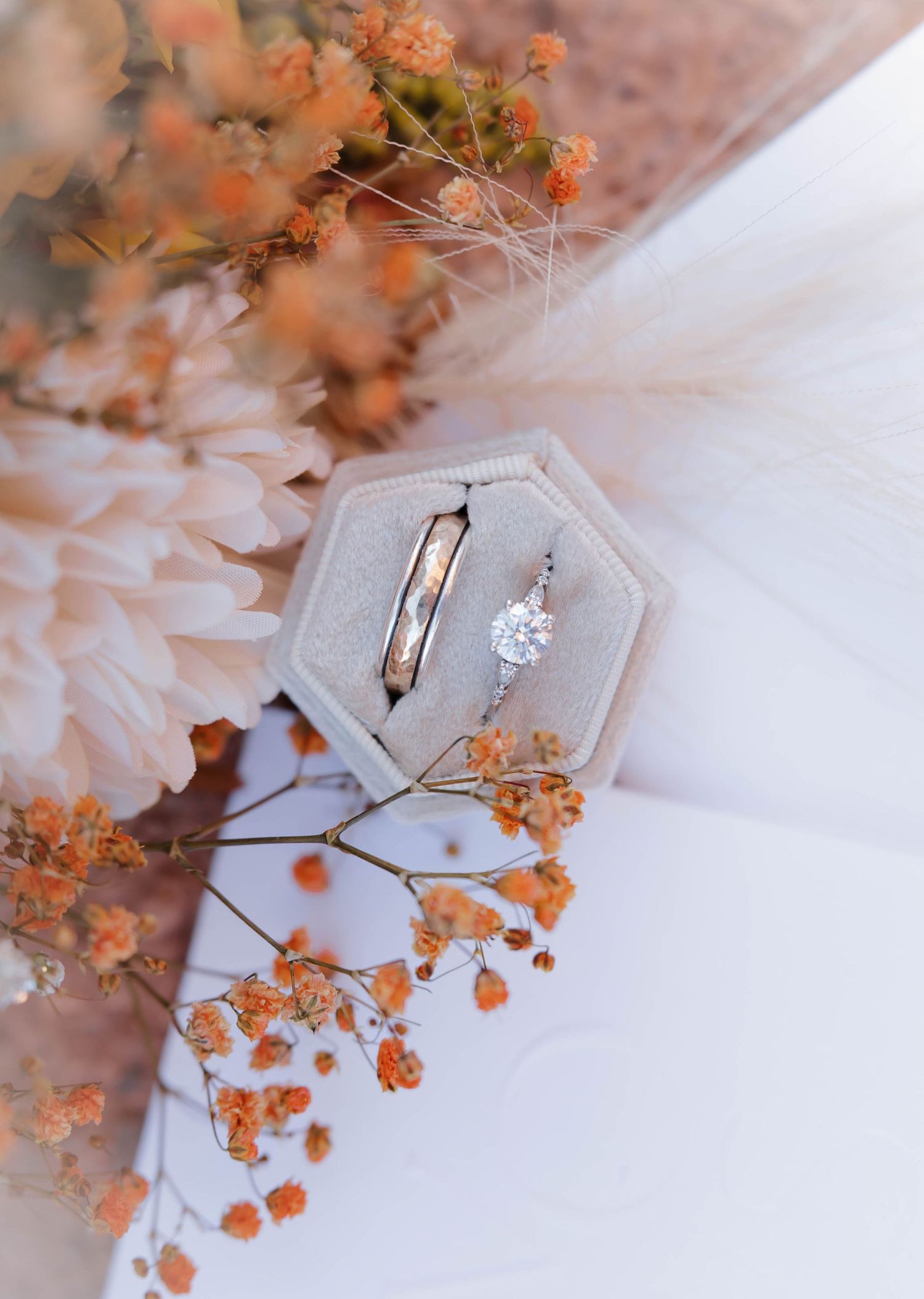 A hexagonal silver engagement ring box with delicate orange dried flowers arranged artistically against white fabric.