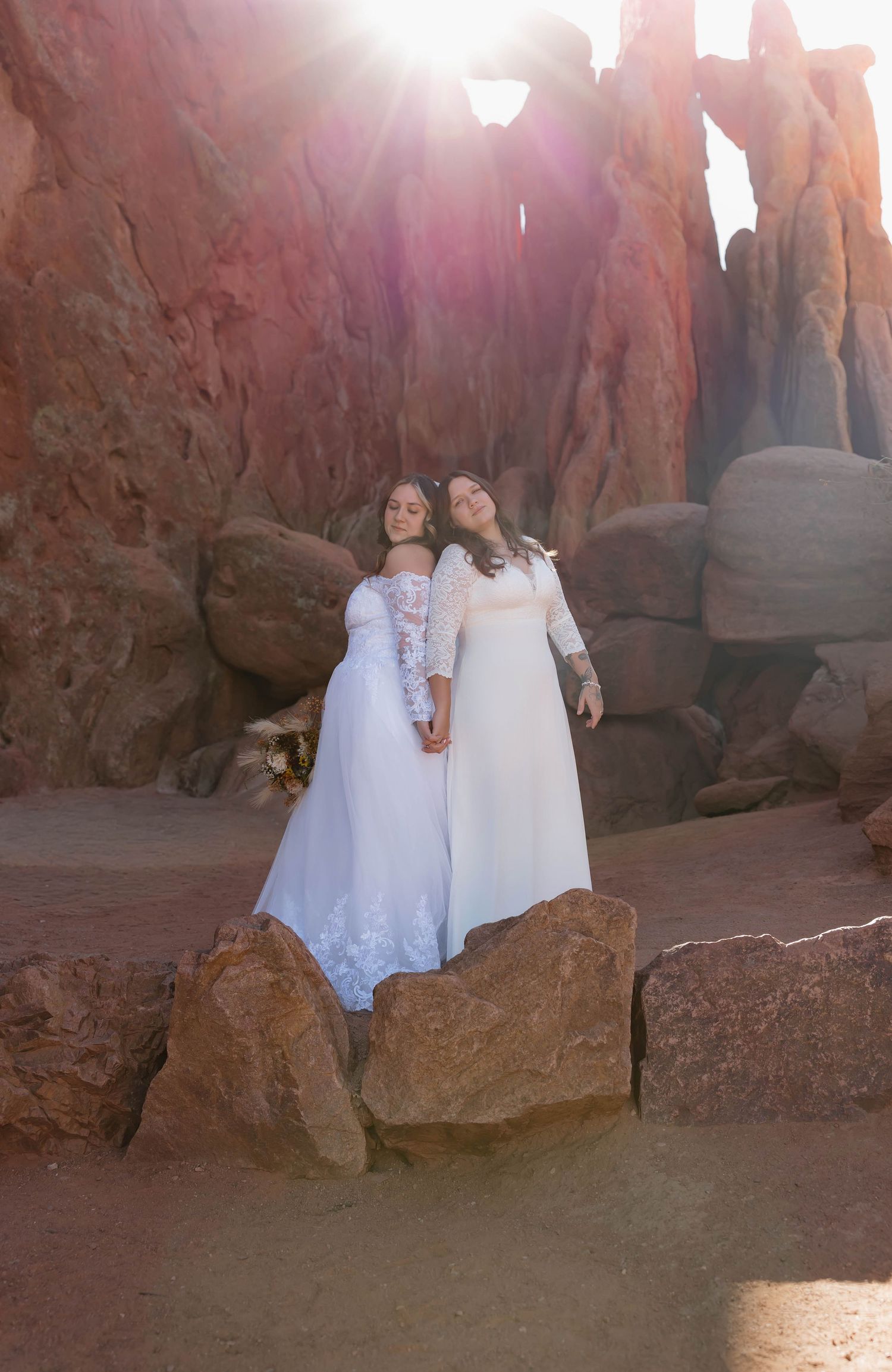 Two people in white dresses sit together on red rock formations in a desert landscape at sunset.