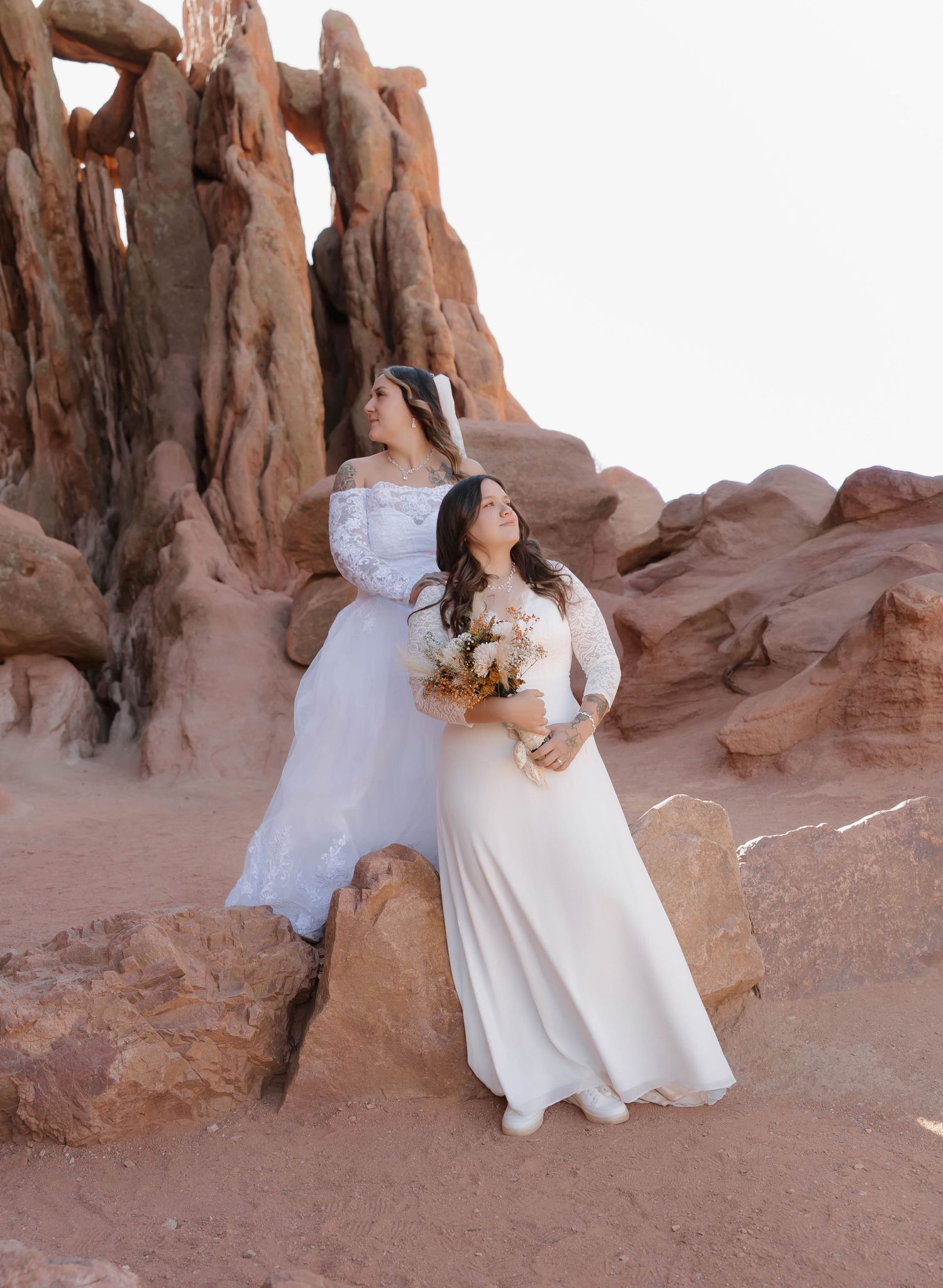 A couple in wedding attire poses together against dramatic red rock formations in a desert landscape.
