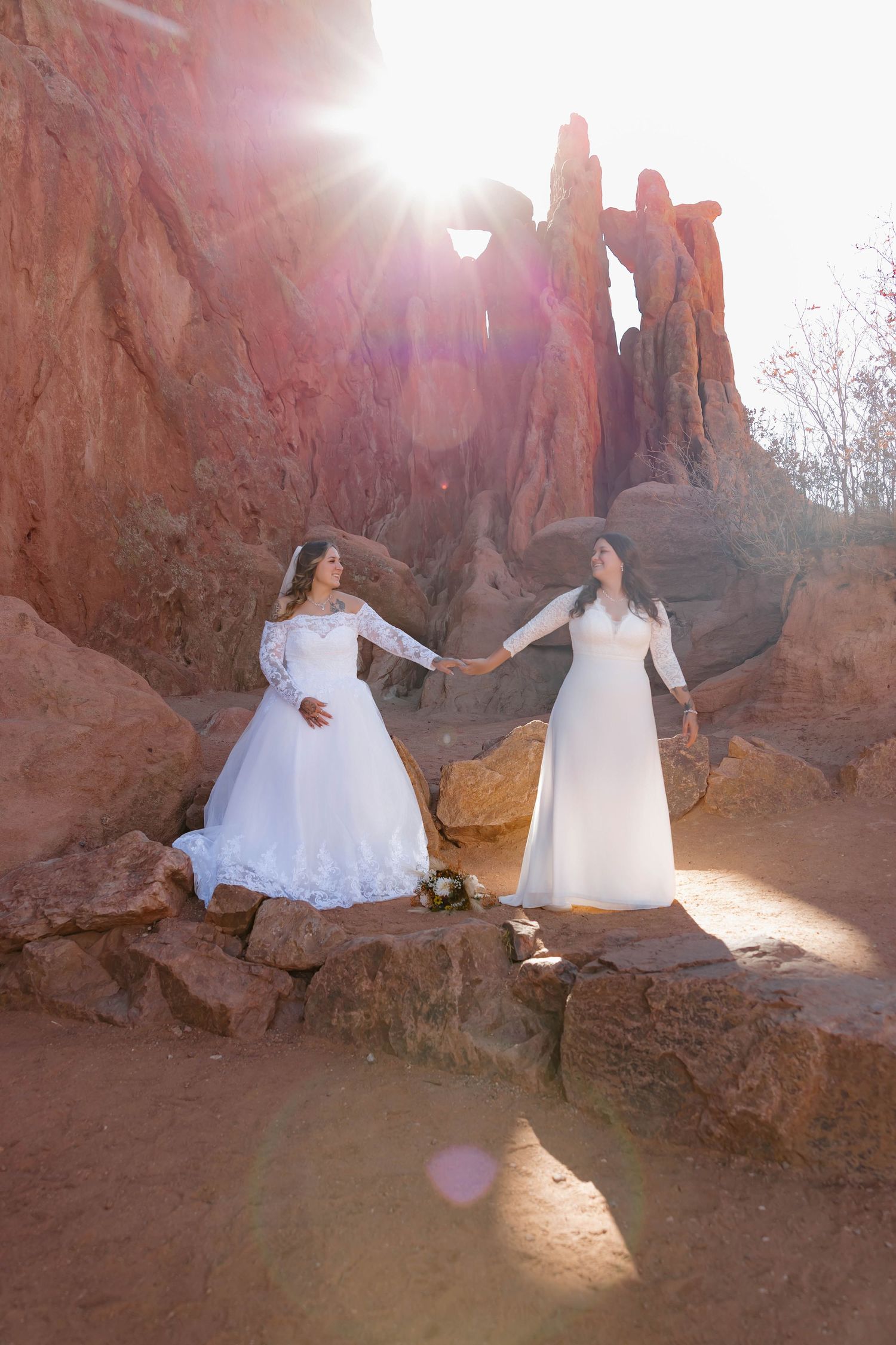Two people in flowing white dresses pose together against red rock formations in a desert landscape at sunset.