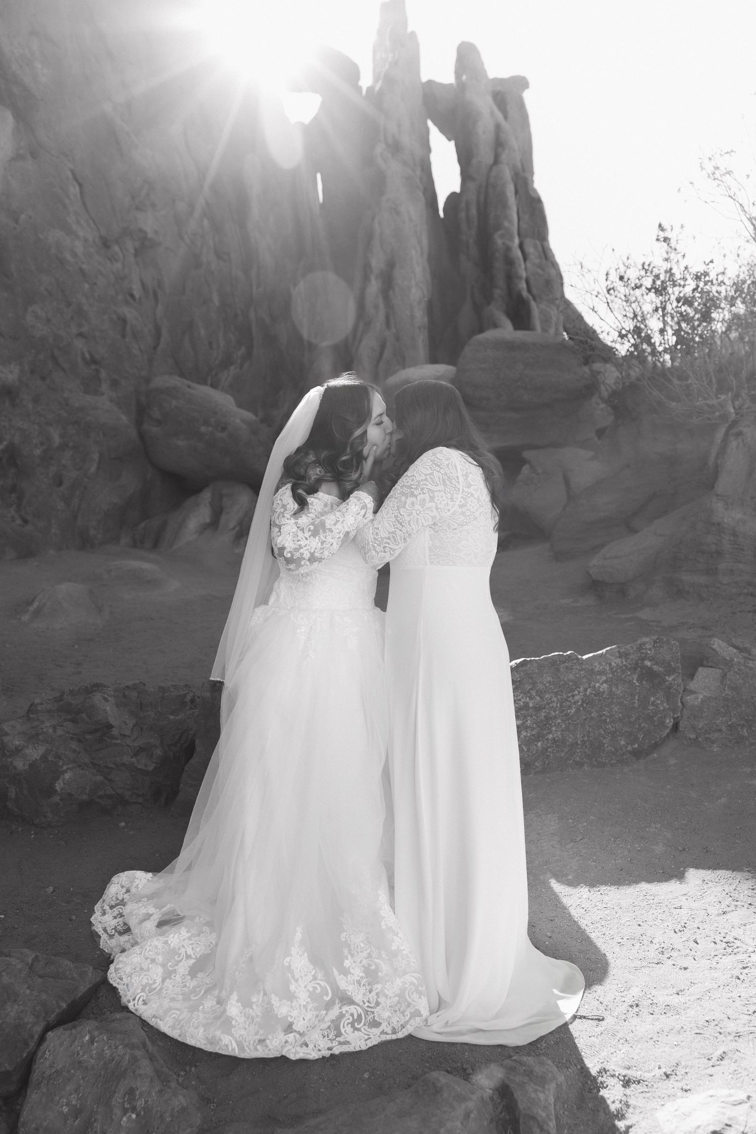 A romantic black and white wedding photo showing two people in white dresses embracing on rocky terrain at sunset.