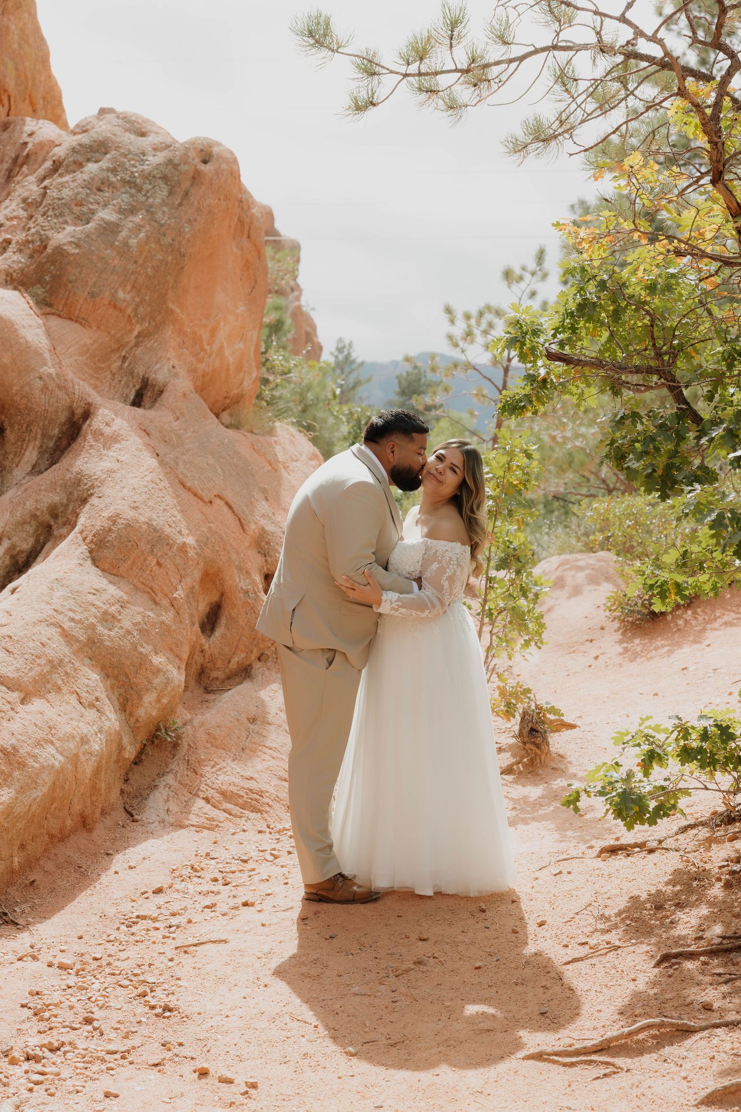 A couple in wedding attire shares a romantic moment among red rock formations on a desert trail.