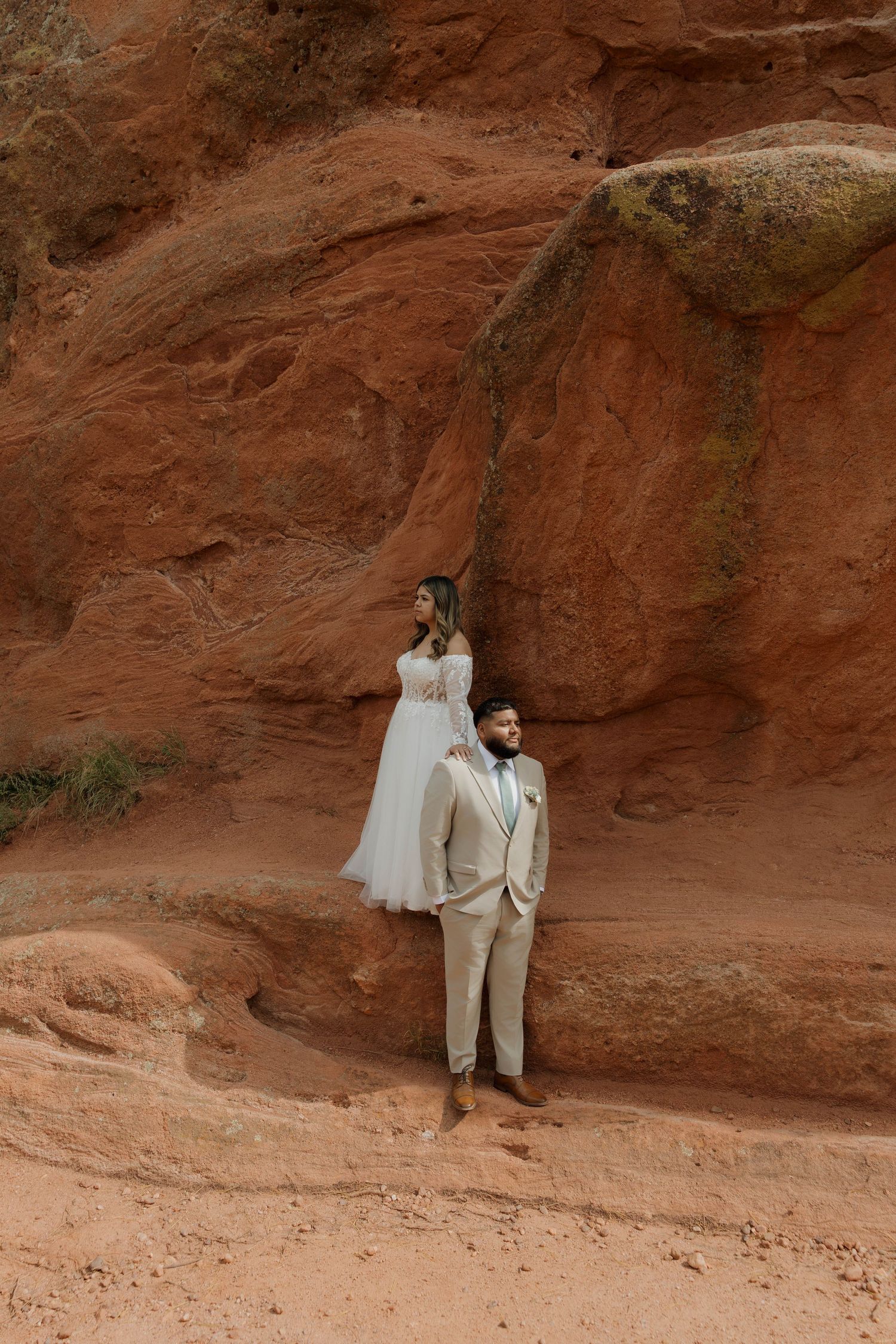 A couple poses against red rock formations in a desert landscape wearing light colored formal attire.