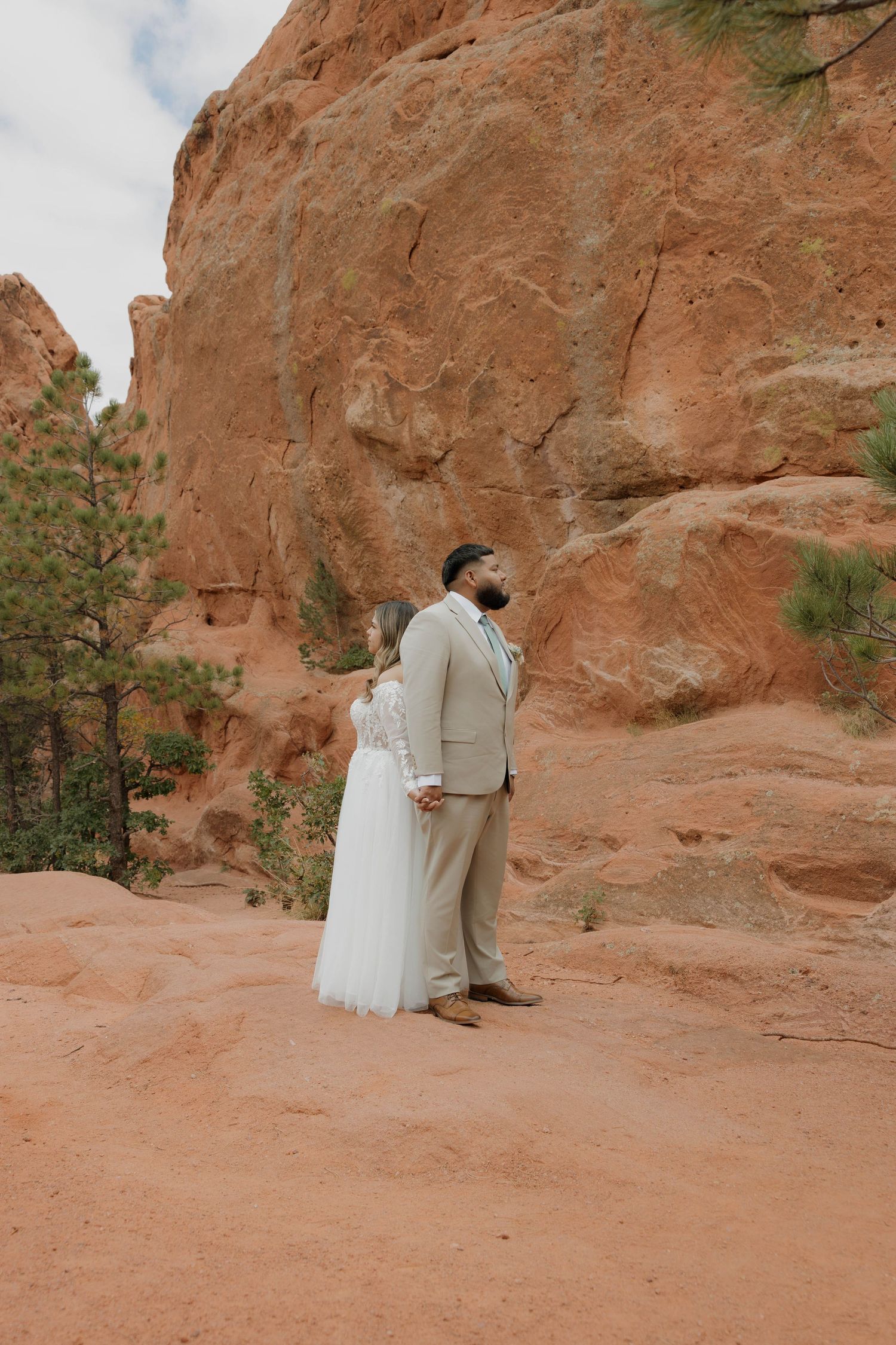 A couple in wedding attire embraces against a stunning red rock formation in a desert landscape.