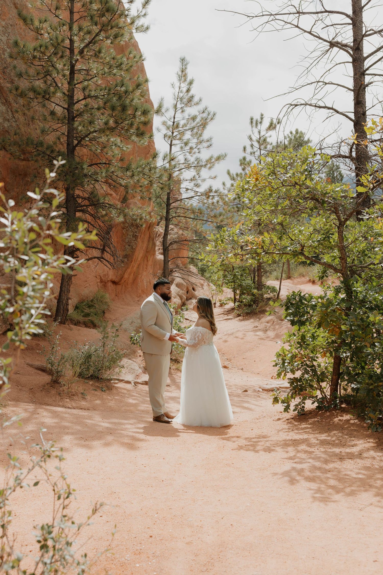 A wedding couple walks along a sandy trail surrounded by pine trees in a natural outdoor setting.