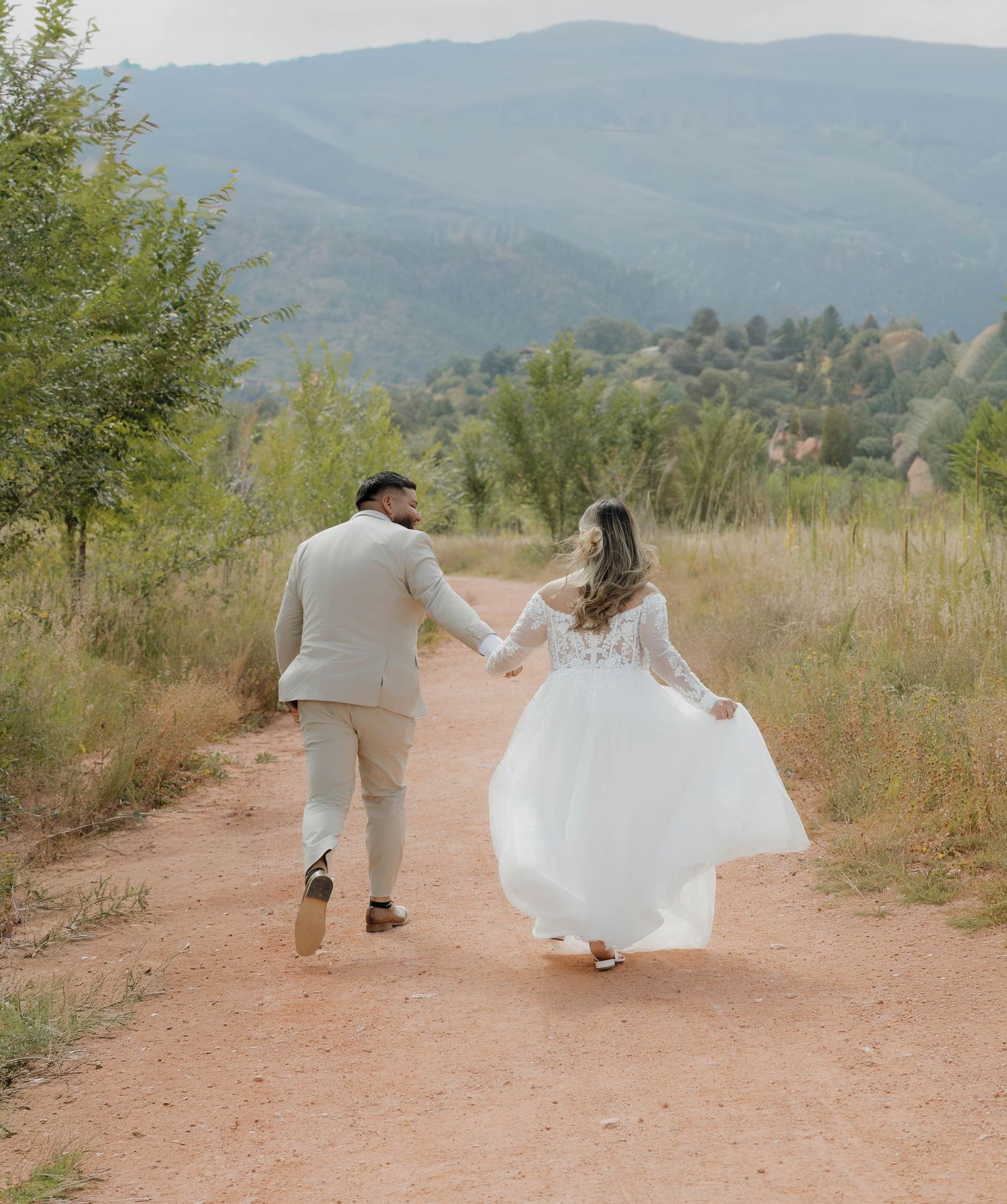 A couple in wedding attire walks along a scenic dirt path with mountains in the background.