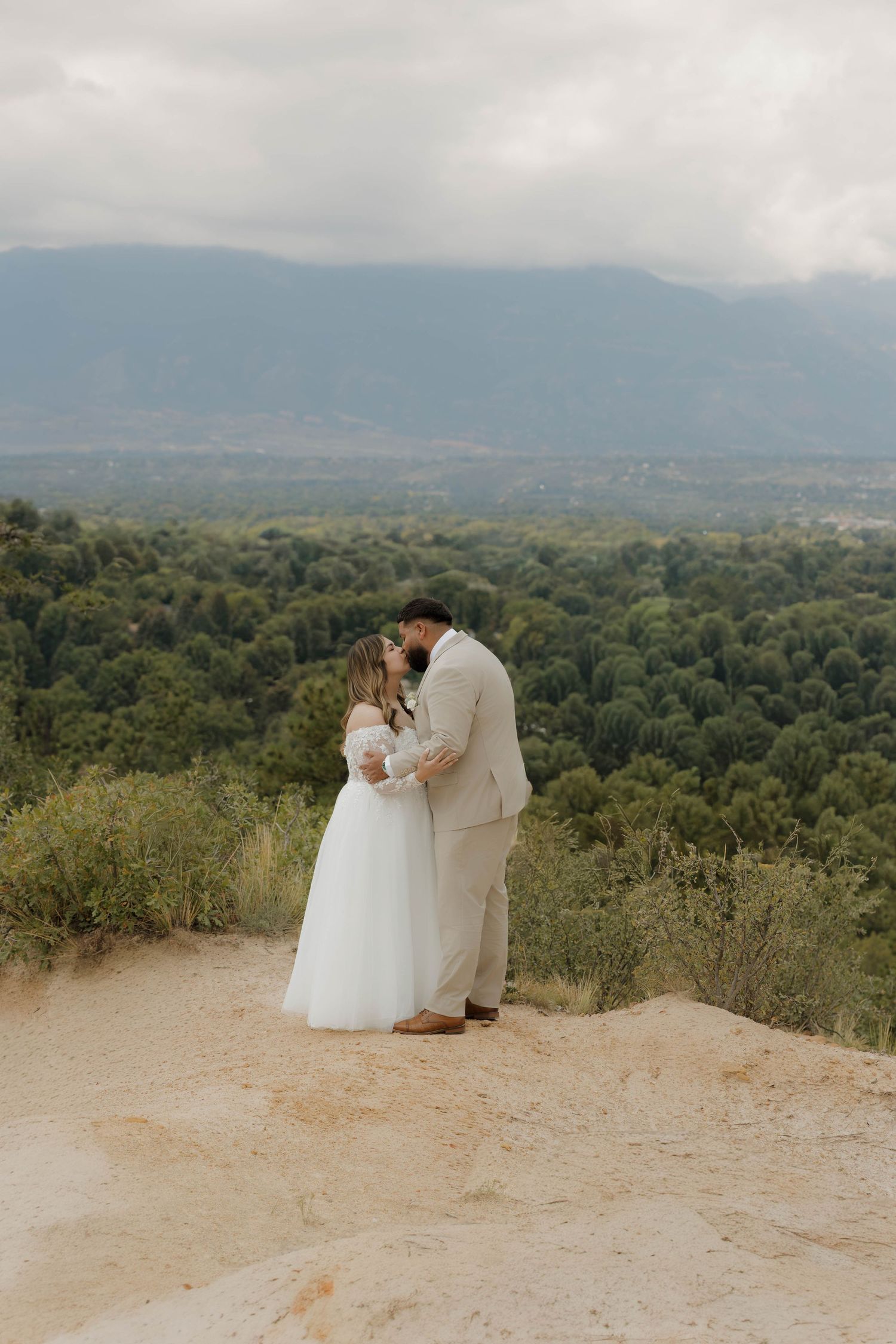 A romantic couple in wedding attire embrace on a scenic mountain overlook with rolling hills and forests in the background.