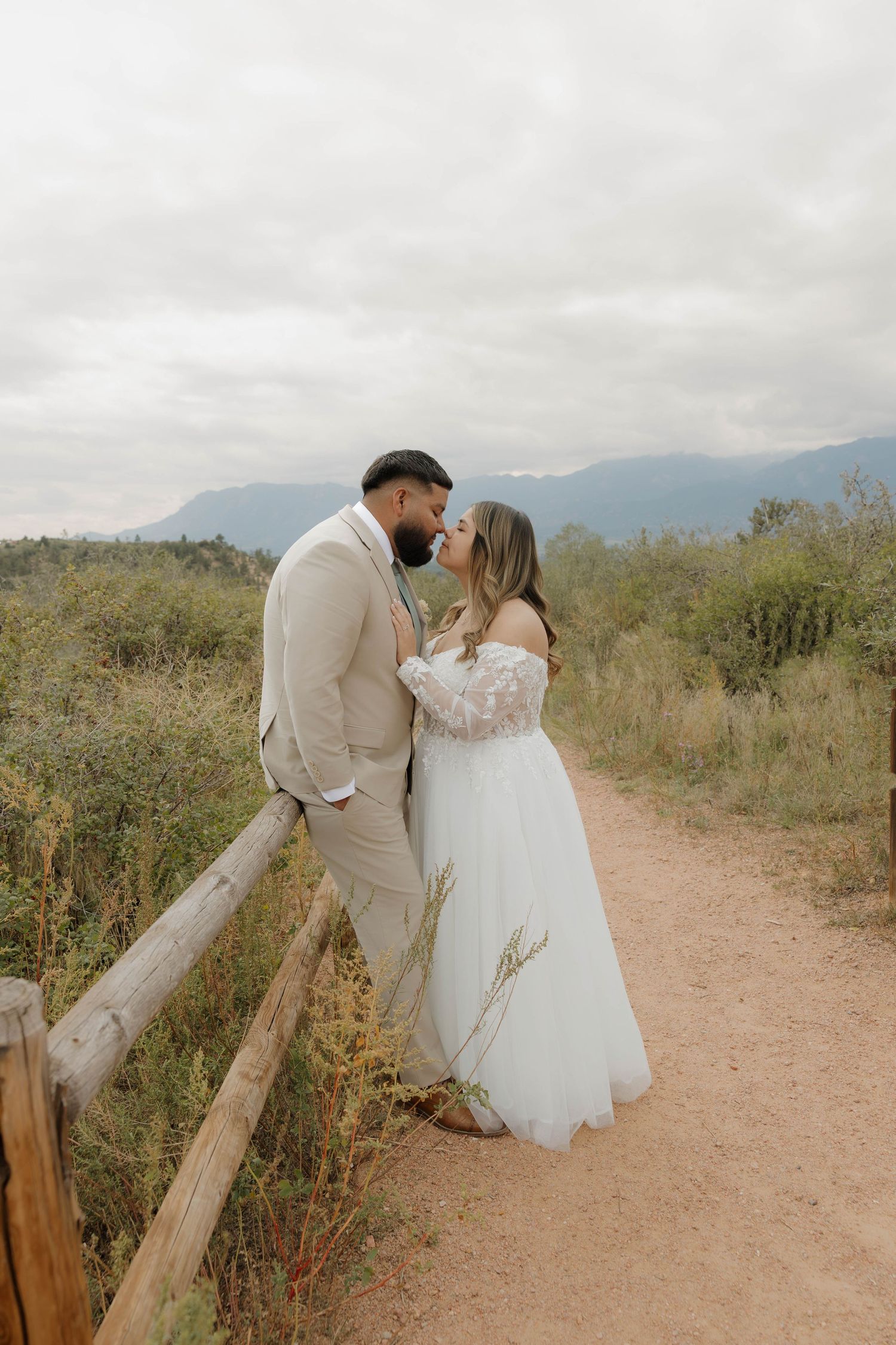 A romantic wedding couple share a tender moment on a dirt trail with mountains and cloudy skies in the background.