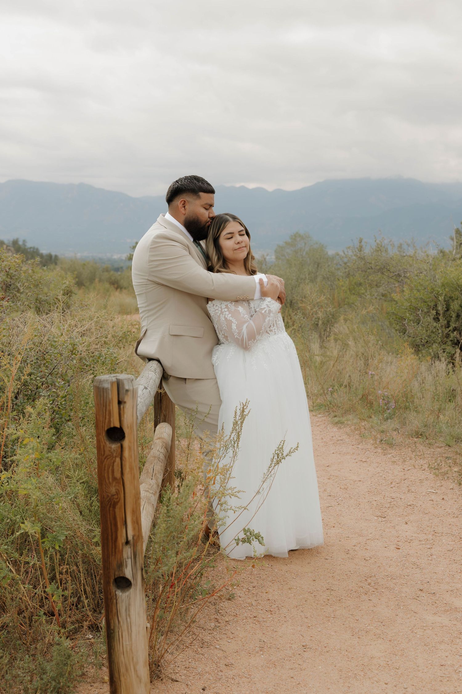 A newlywed couple embraces on a rustic mountain path with a wooden fence and natural landscape surrounding them.