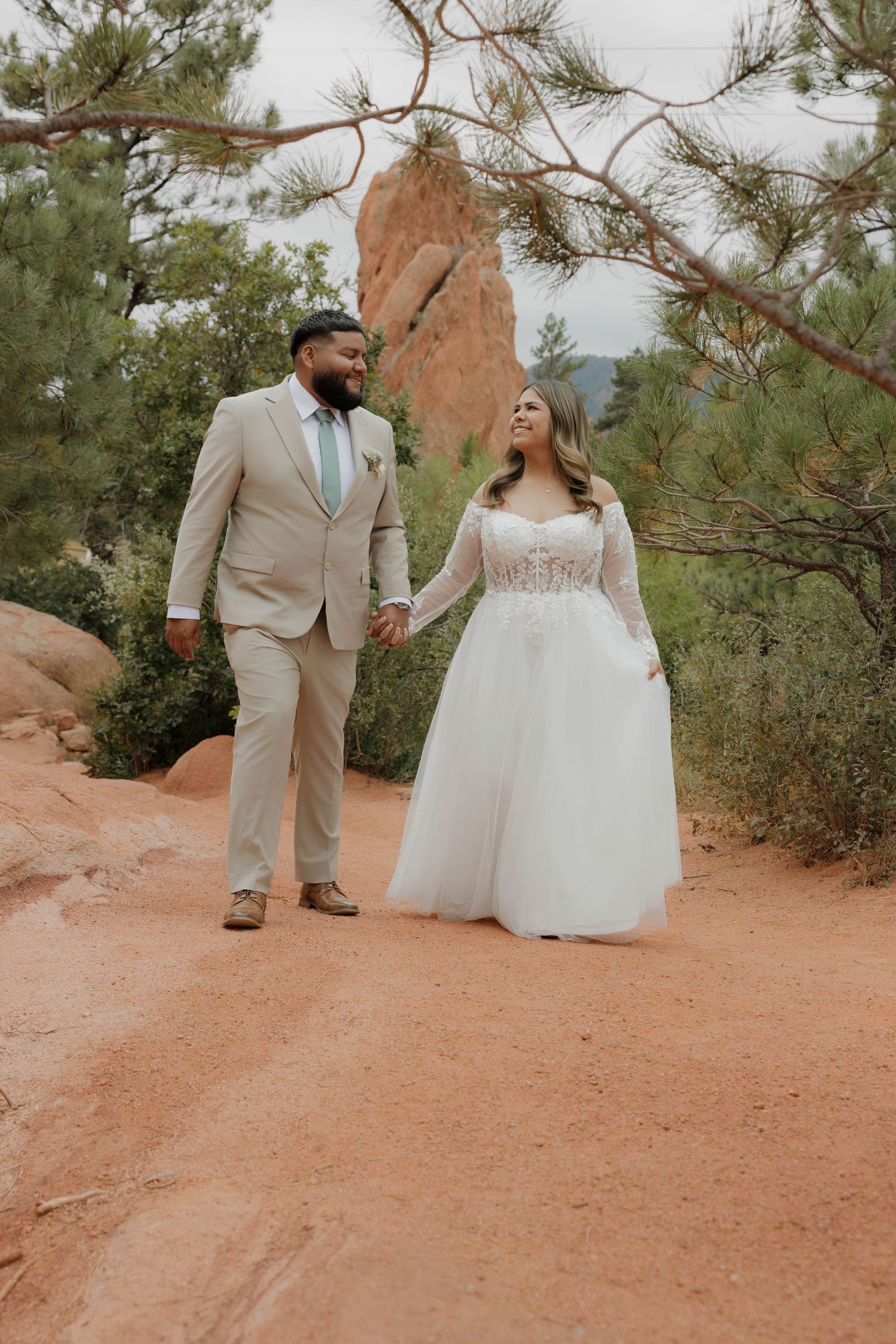 Newlywed couple walking along a red dirt path surrounded by pine trees in a romantic outdoor setting.