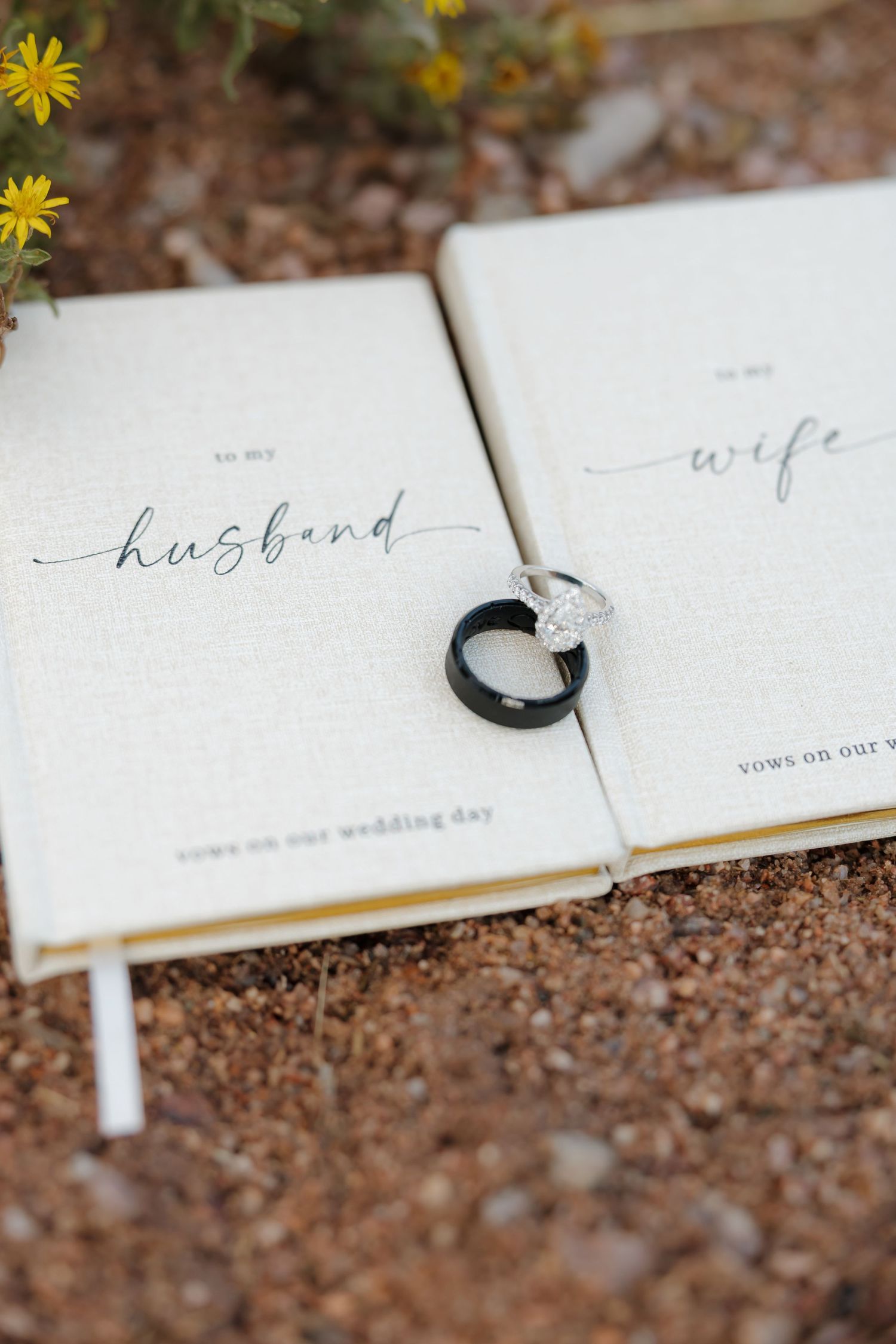 Wedding rings rest on open vow books labeled 'husband' and 'wife' with yellow flowers in the background.
