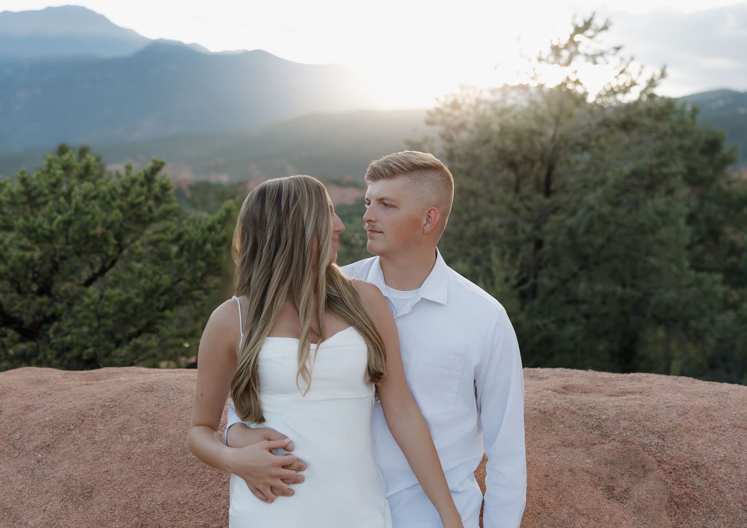 A couple in white outfits embraces while standing on a mountain overlook during golden hour.