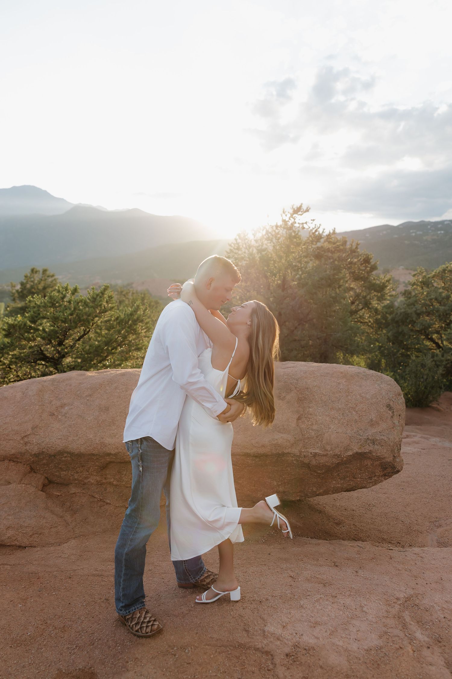 A couple shares a romantic sunset moment on a rocky overlook with mountains in the background.