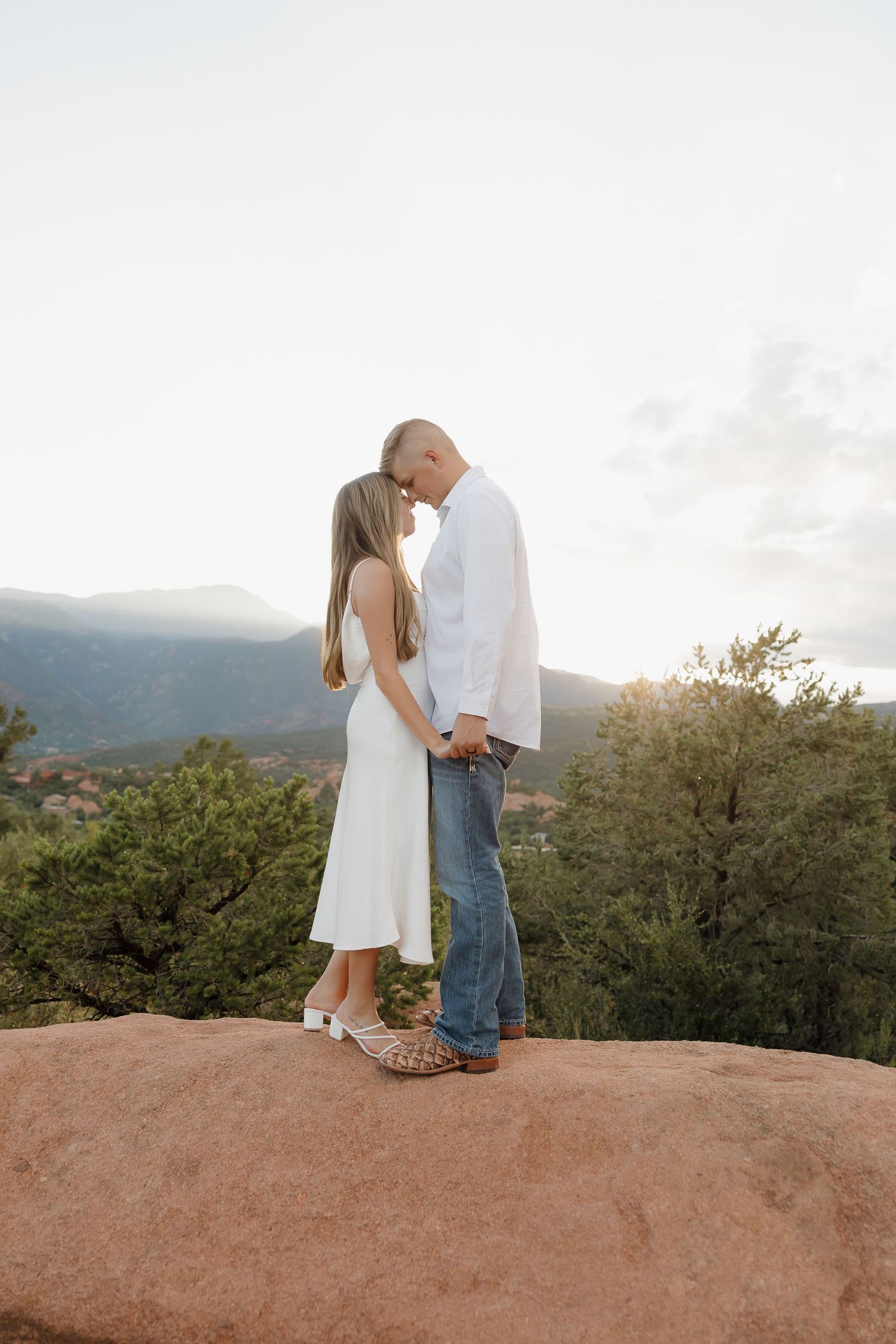A romantic couple stands intimately on a red rock overlook at sunset with mountain views in the background.