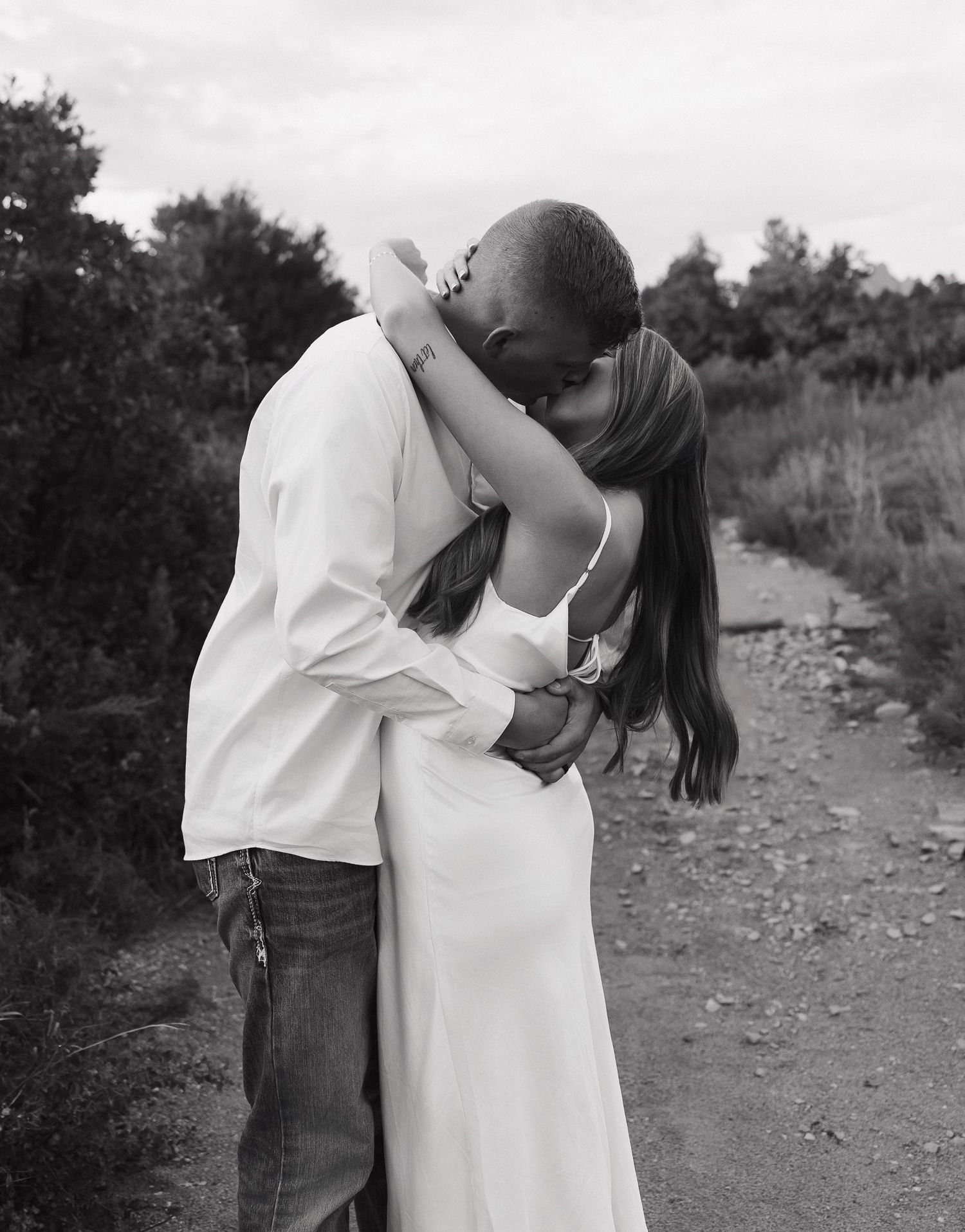 A romantic couple embraces on a dirt path in a black and white photograph surrounded by trees and nature.