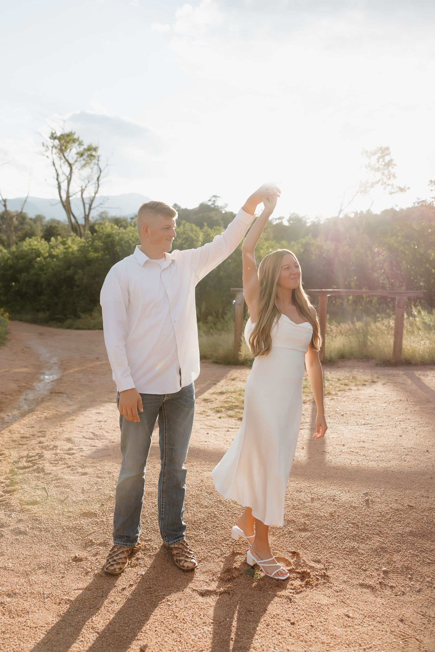 A couple dances together on a dusty path at sunset while wearing casual white outfits in a desert setting.