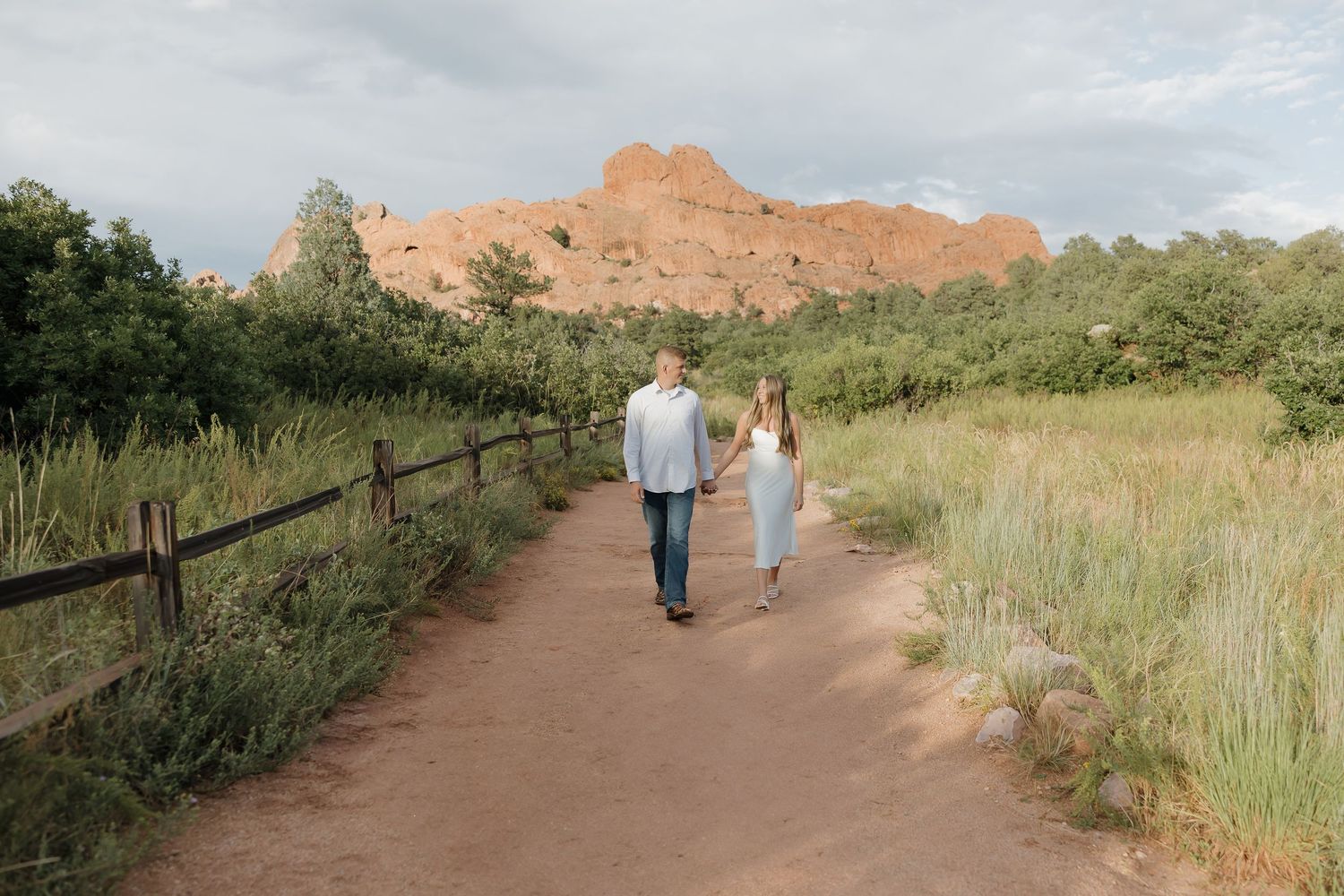 A scenic dirt trail winds through green grasslands with red rock formations visible in the background under cloudy skies.