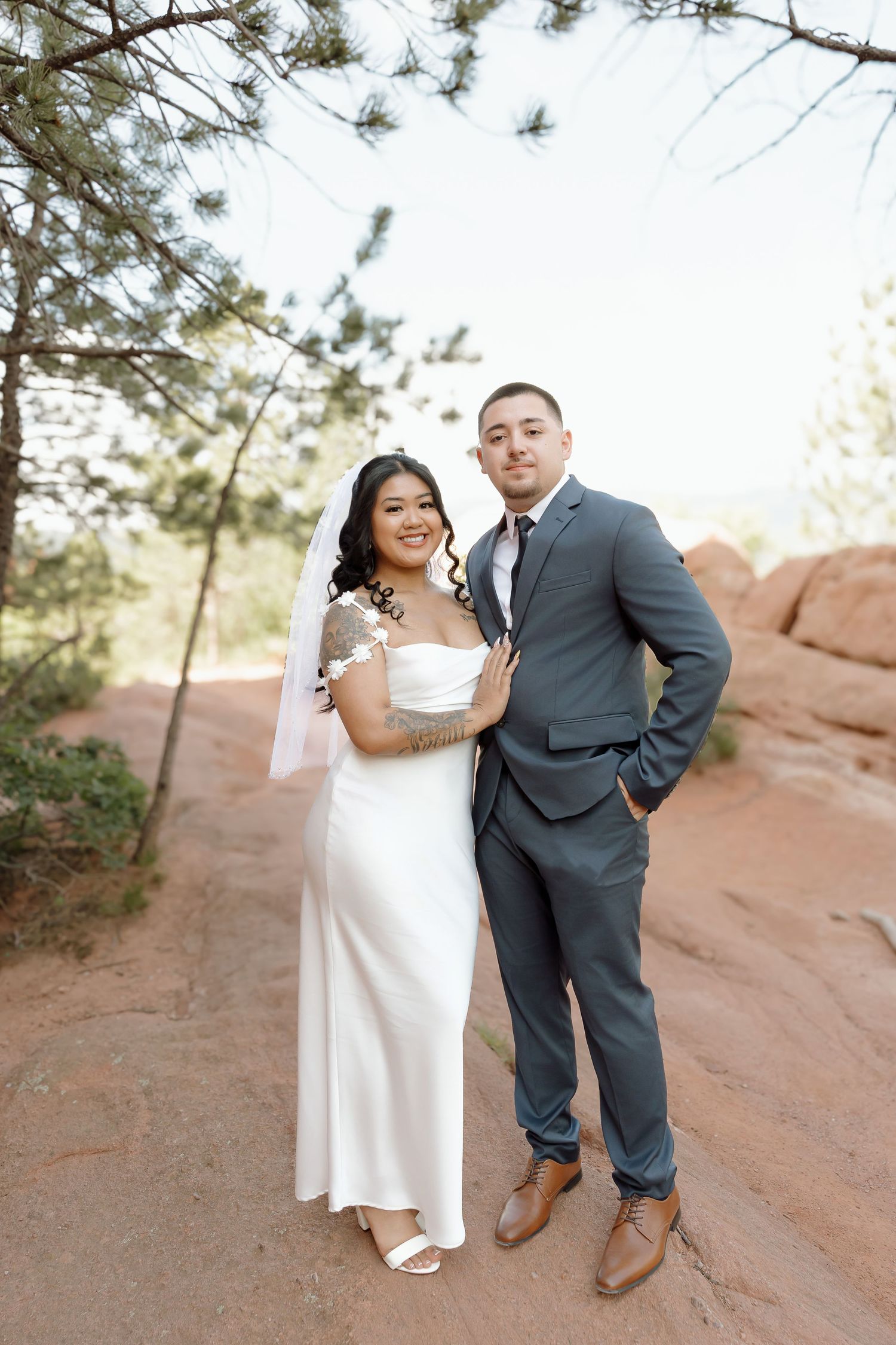 A newlywed couple poses together on a red rock trail surrounded by pine trees in an outdoor setting.