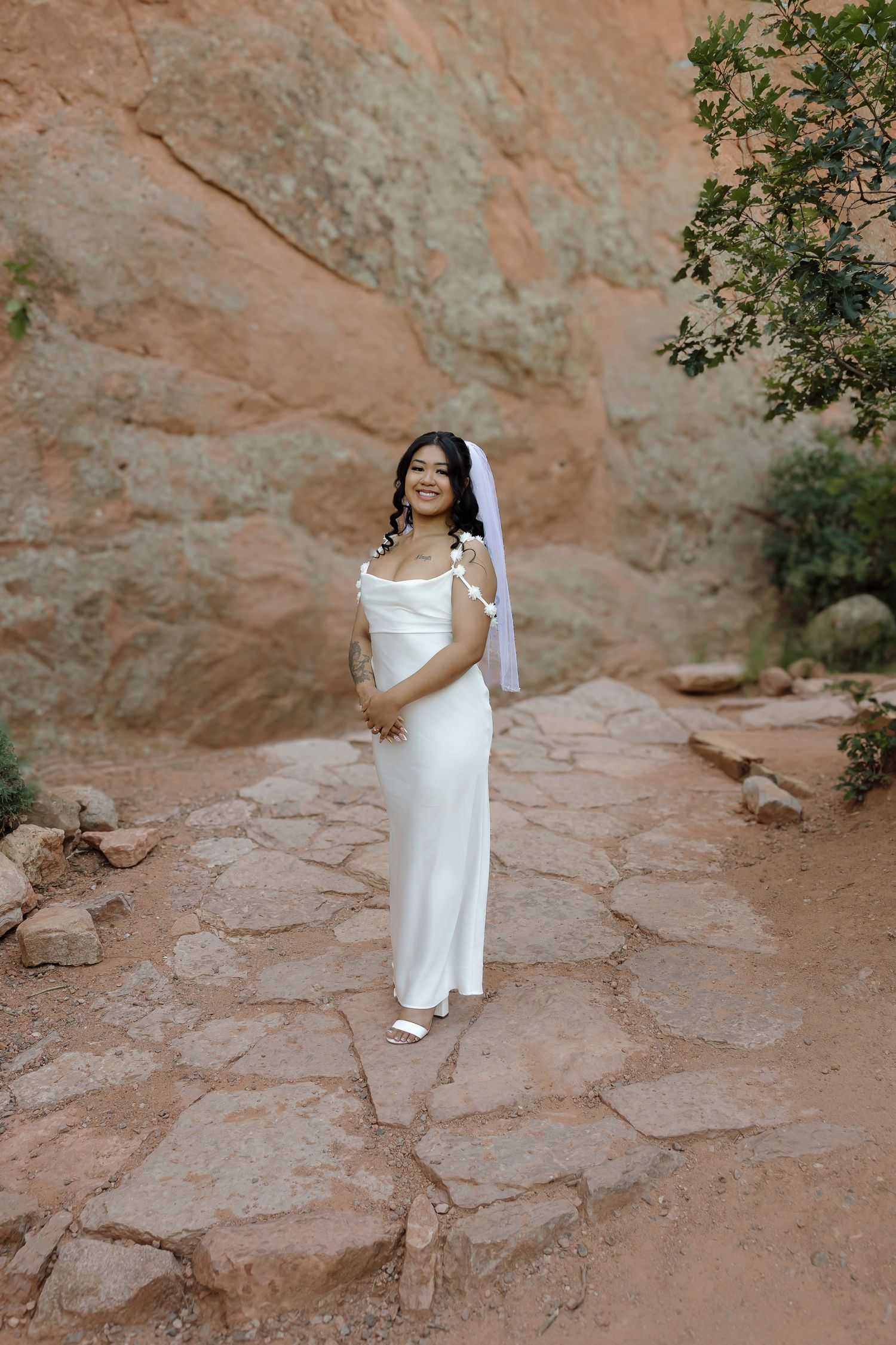 Bride in elegant white dress poses on red rock formations in natural outdoor setting.
