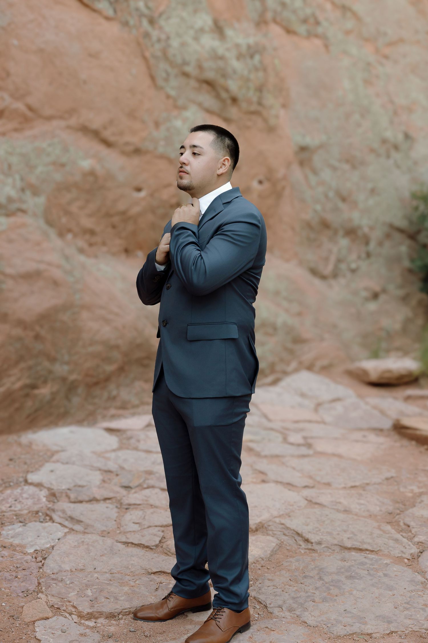 A groom in a navy suit poses thoughtfully against a red rock formation backdrop.